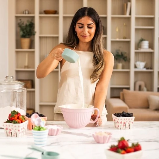 Woman baking in a kitchen, pouring liquid into a mixing bowl with berries on the counter.
