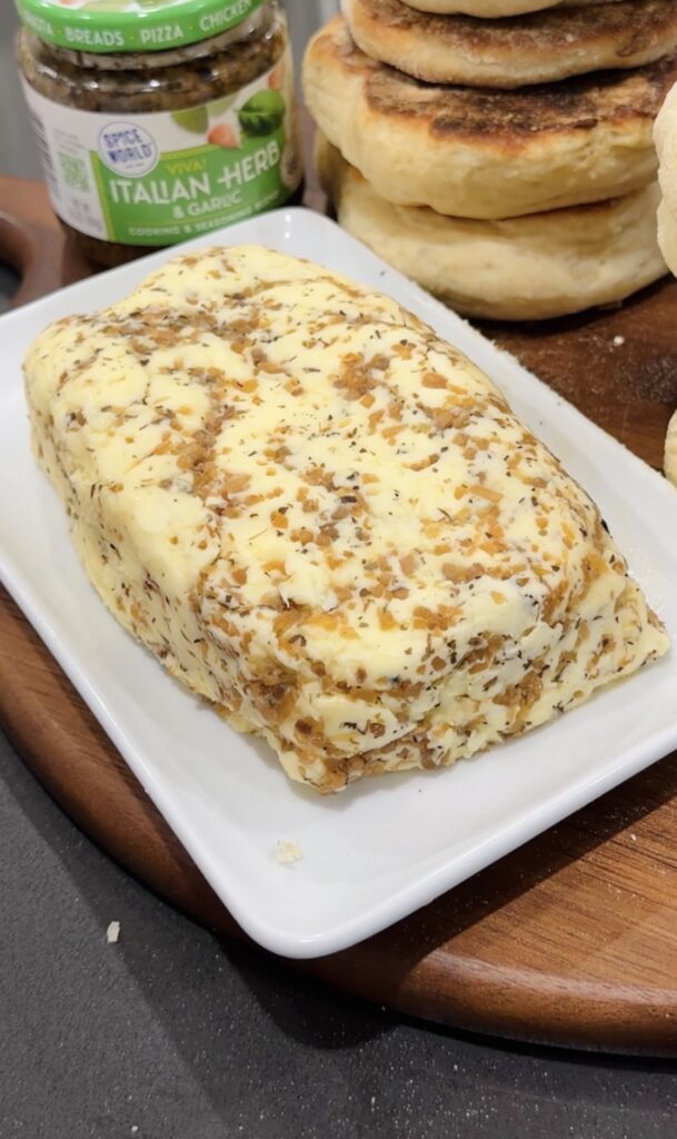 Block of garlic and herb compound butter on a white plate, next to English muffins and a jar of Italian herbs.