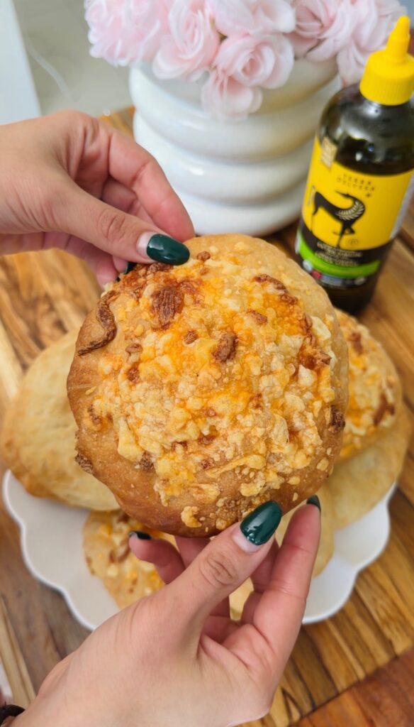 Homemade focaccia rolls topped with cheese, held by hands with dark green nails. Olive oil bottle in background.